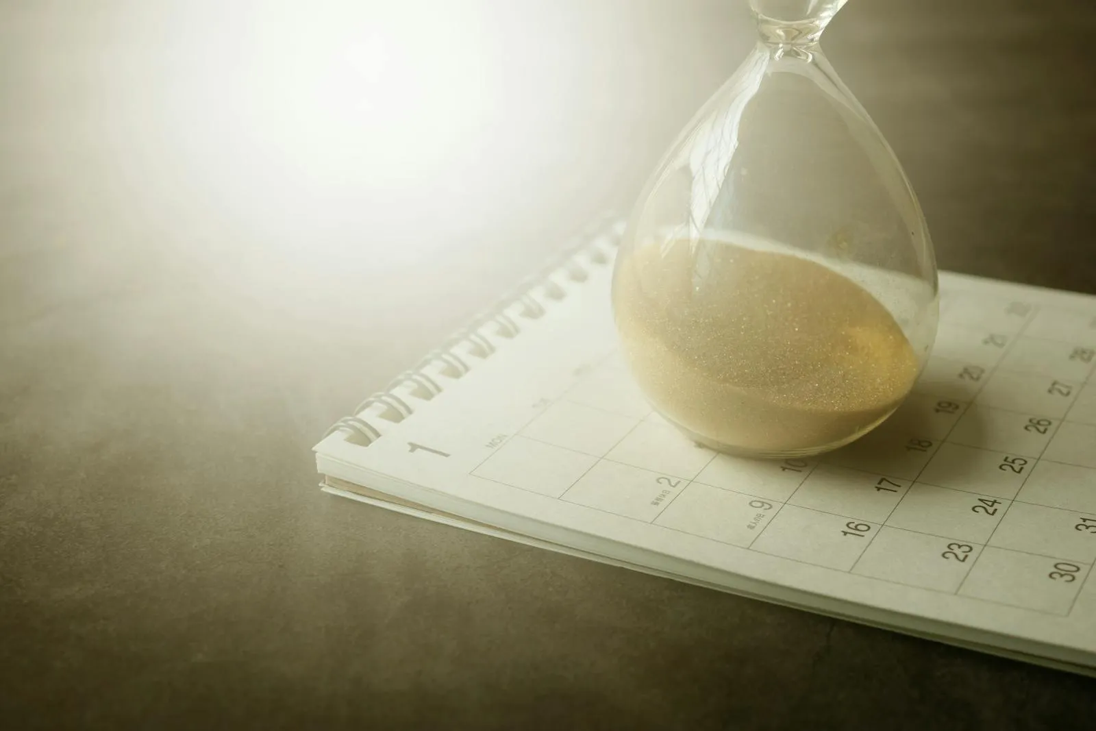 Sablier au sable beige posé sur un calendrier mensuel ouvert sur fond gris